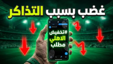 هاتف ذكي يعرض هاشتاج مطالبة بتخفيض التذاكر بخلفية لملعب كرة قدم مضاء باللون الأخضر.
